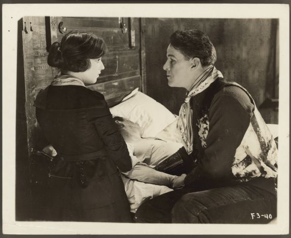 Helen Russell and William Russell appear in a still from the 1920 film <i>Shod with Fire</i>.  Ferguson kneels next a man in a bed while Russell sits next to the man on the bed.  The two look intently at each other.